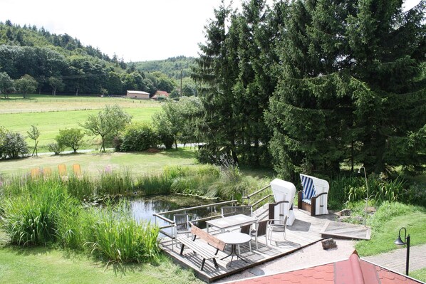 Eine Holzterrasse mit Gartenmöbeln, ein kleiner Teich und Blick auf einen bewaldeten Hang.