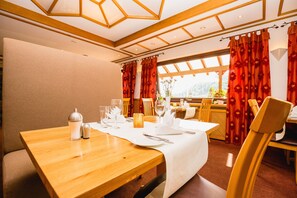 Ein Essbereich mit einem Holztisch, gedeckt für zwei Personen, einer Blumenvase und Blick ins Freie durch große Fenster.