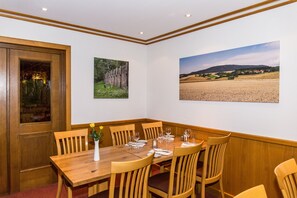 Ein Esszimmer mit einem Holztisch, gedeckt für sechs Personen, umgeben von Holzstühlen. An den Wänden hängen zwei gerahmte Bilder, eines zeigt eine Waldszene, das andere eine ländliche Landschaft.