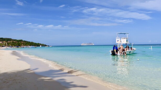 A beach with clear turquoise water, a boat with people, and a cruise ship in the distance. - La Placita Inn (Roatan)