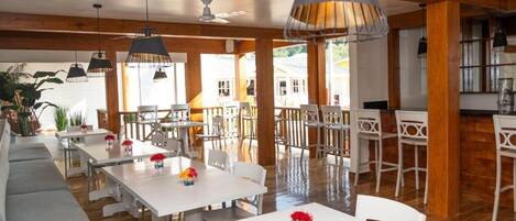 Un vaste espace dédié à la salle à manger, avec des tables et des chaises blanches, des poutres en bois au plafond et des luminaires suspendus.