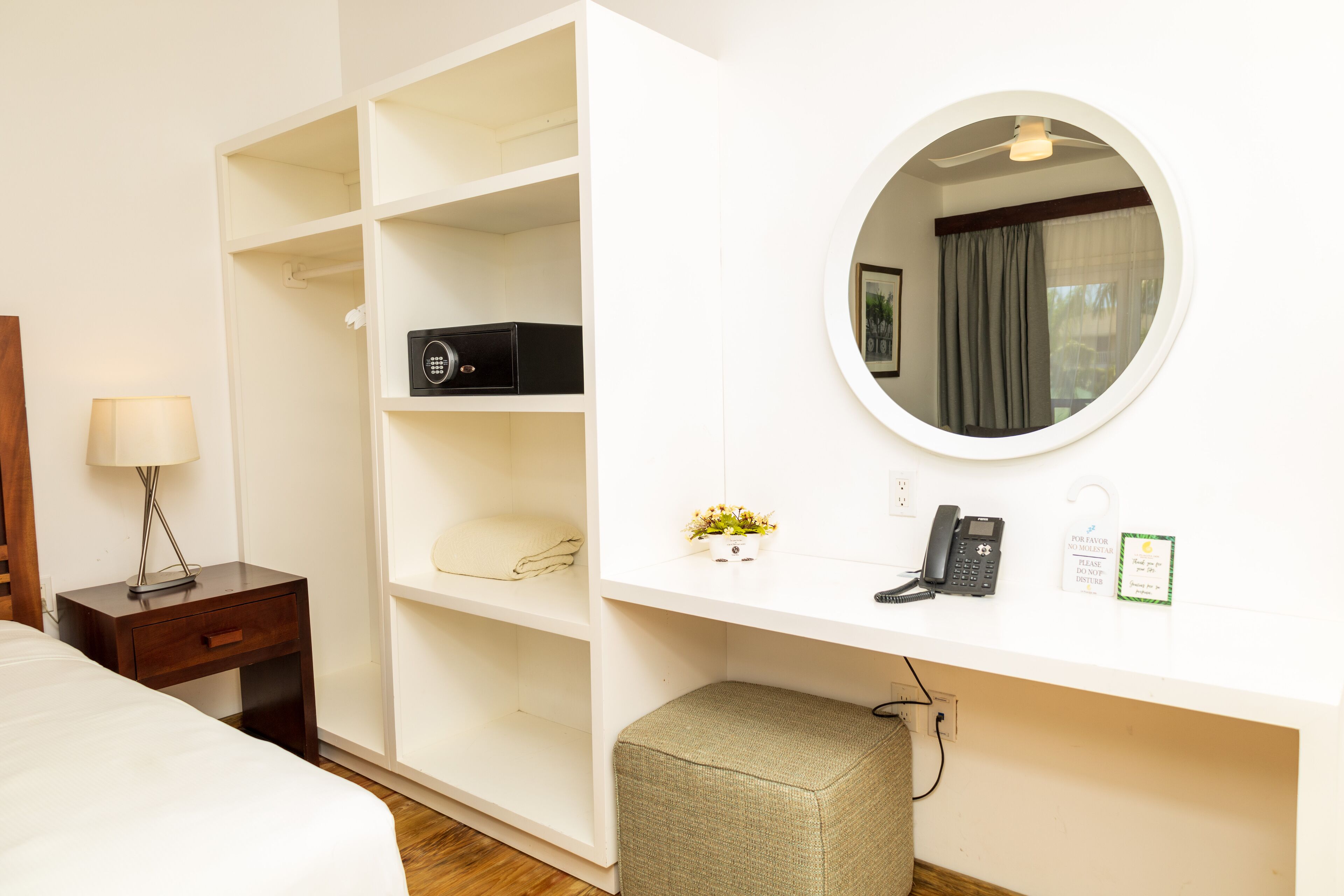A hotel room with a bed, a nightstand with a lamp, a wardrobe, a desk with a telephone, a mirror, and a small stool.