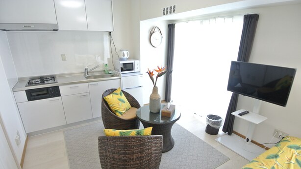 A modern kitchen with white cabinets, a microwave, and a stove. There is a dining area with wicker furniture and a small table. A flat-screen TV is mounted on the wall, and a window with curtains is visible. - Kukuru House 3 (Naha)