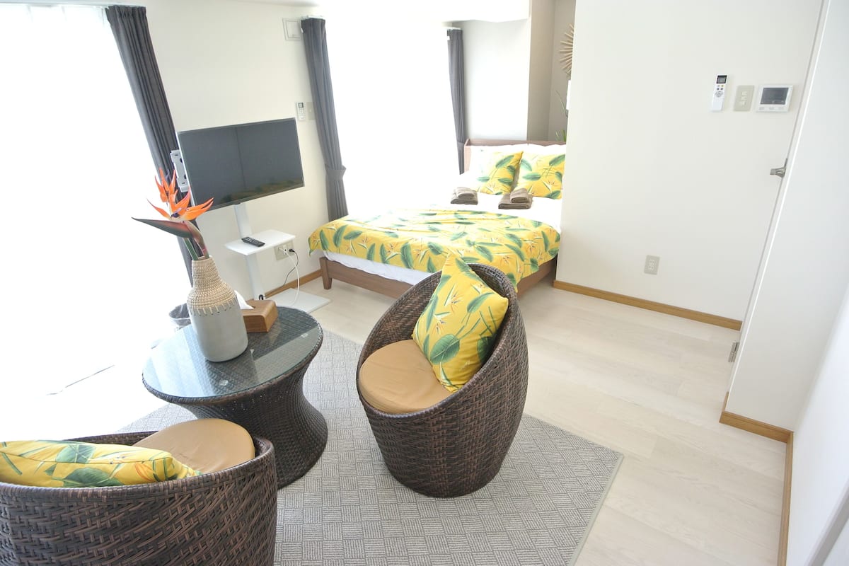 Habitación de hotel con cama, dos sillas de mimbre, una mesita con un florero, televisor de pantalla plana y ventana con cortinas.