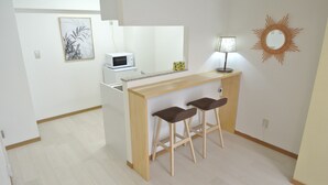 A small kitchenette with a microwave, a coffee maker, and a lamp on a wooden counter. There are two stools and a framed picture on the wall. - Kukuru House 3 (Naha)