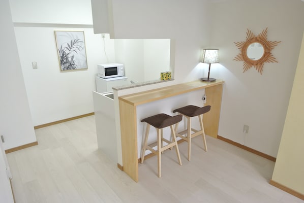 A small kitchenette with a microwave, a coffee maker, and a lamp on a wooden counter. There are two stools and a framed picture on the wall.