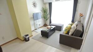 A living room with a sofa, coffee table, TV, potted plant, and a trash bin. - Kukuru House 3 (Naha)