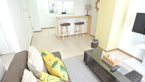 A modern living room with a sofa, a glass coffee table, and a kitchen area with a bar stool. - Kukuru House 3 (Naha)