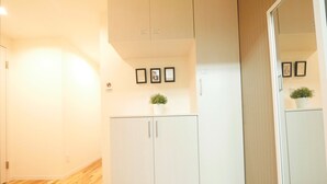 A hallway with a wooden floor, white walls, and a white wardrobe with a small potted plant on top. There are framed pictures on the wall and a mirror on the right.