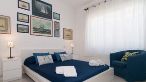 Ein ordentlich eingerichtetes Schlafzimmer mit einem großen Bett, zwei Nachttischen, einem Sessel und einem Fenster mit Vorhängen.