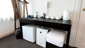 Ett kompakt hotellrum med ett litet skrivbord, ett minikylskåp, en liten kylbox, en vit bänk, en hårtork, en vattenkokare, en kaffebryggare och en väckarklocka på väggen.
