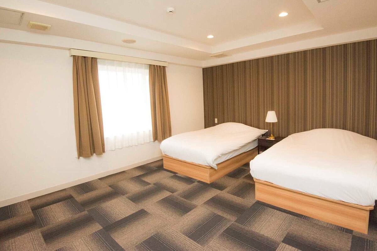 Habitación de hotel con dos camas, una ventana con cortinas, una lámpara de pared y una alfombra estampada.