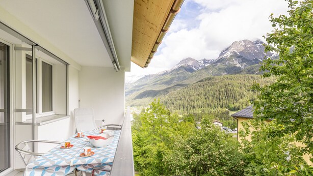 Ein Balkon mit Tisch und Stühlen, mit Blick auf eine gebirgige Landschaft mit Bäumen und einem Gebäude in der Ferne.