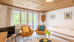 Ein modernes Zimmer mit Holzboden, einem Fernseher, zwei Stühlen, einer Couch und einem Fenster mit Blick auf Grünflächen.