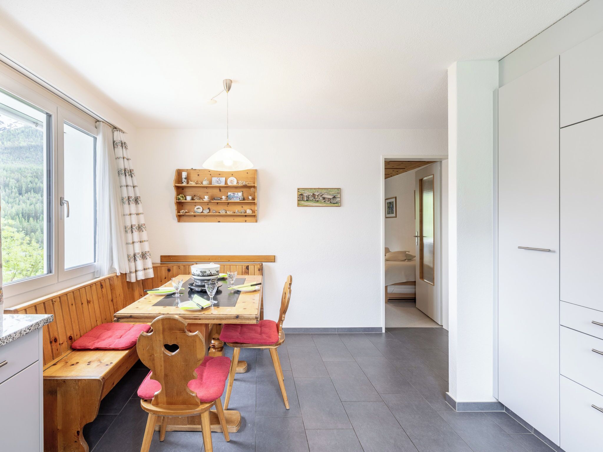 A dining area with a wooden table and chairs, a pendant light, and a shelf with kitchenware.