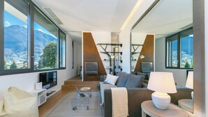 Un salon moderne avec un canapé, une table basse et une lampe. Une grande fenêtre offre une vue sur les montagnes et la ville.
