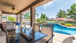 Eine Außenpatio mit Glastisch, Stühlen, Deckenventilator und Blick auf Pool und Grünflächen.