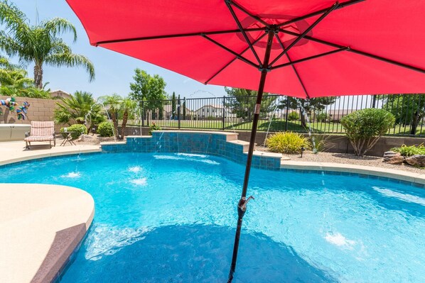 Une piscine avec un parasol rouge, entourée de palmiers et d’une clôture.