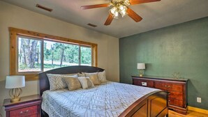 Un dormitorio con cama, mesita de noche, cómoda, ventilador de techo y ventana con vista a árboles.