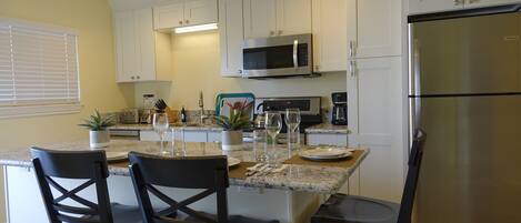 A kitchen with white cabinets, a stainless steel refrigerator, a microwave, and a coffee maker. There are two chairs and a table set with plates, glasses, and cutlery.