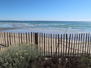 Een strand met een houten omheining op de voorgrond en mensen in het water achterin.
