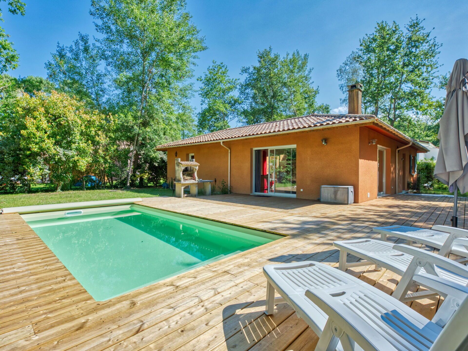 Una piscina con muelle de madera, zona de barbacoa y terraza techada con sillones de descanso.