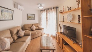 A living room with a beige sofa, a glass coffee table, a wooden TV stand, and a flat-screen TV. There are decorative items on the shelves, and a painting is on the wall. - Apartamento Cóncavos  (Zahara de los Atunes)