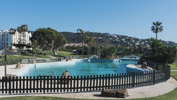 A large outdoor swimming pool with a wooden fence, surrounded by palm trees and a view of a hillside with buildings. - Apartamento Cóncavos  (Zahara de los Atunes)