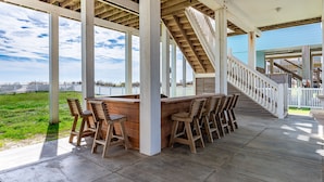 A covered outdoor area with a wooden bar and seating, a staircase, and a view of a fenced property.