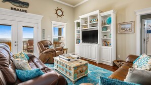 A living room with a leather couch, a coffee table, and a TV stand. There is a bookshelf and a door leading to a balcony.