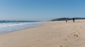 Una amplia playa de arena con olas oceánicas, cielo despejado y un paisaje montañoso en la distancia.