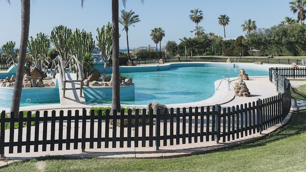 Una piscina con cerca de madera, rodeada de palmeras y rocas.