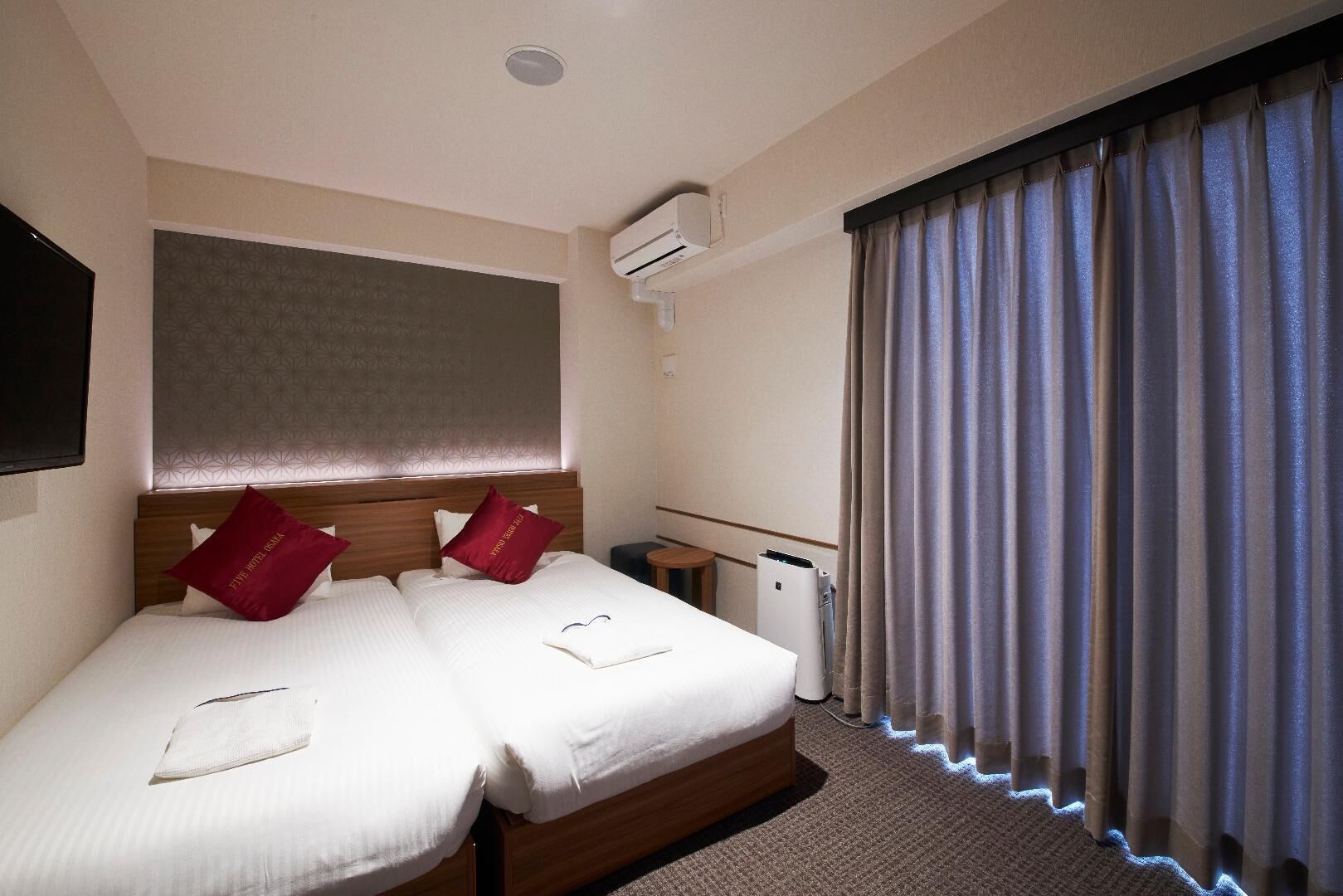 Quarto de hotel com duas camas, cabeceira, ar condicionado, uma pequena mesa e uma janela com cortinas.