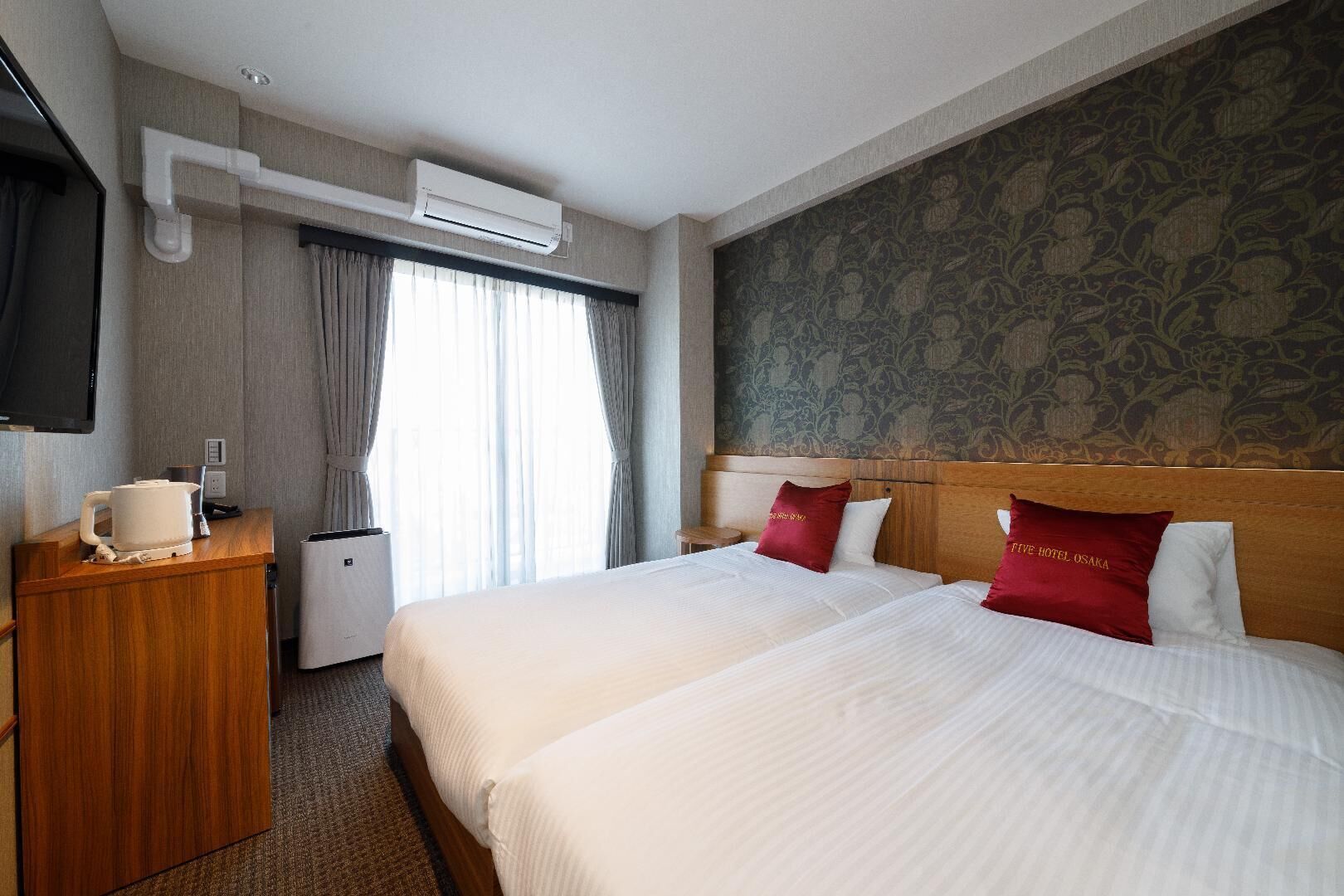 Ein Hotelzimmer mit zwei Betten, einem Holzkopfende, einem Flachbildfernseher, einer Klimaanlage, einem kleinen Tisch mit Wasserkocher und einer Wand mit Musterpapier.