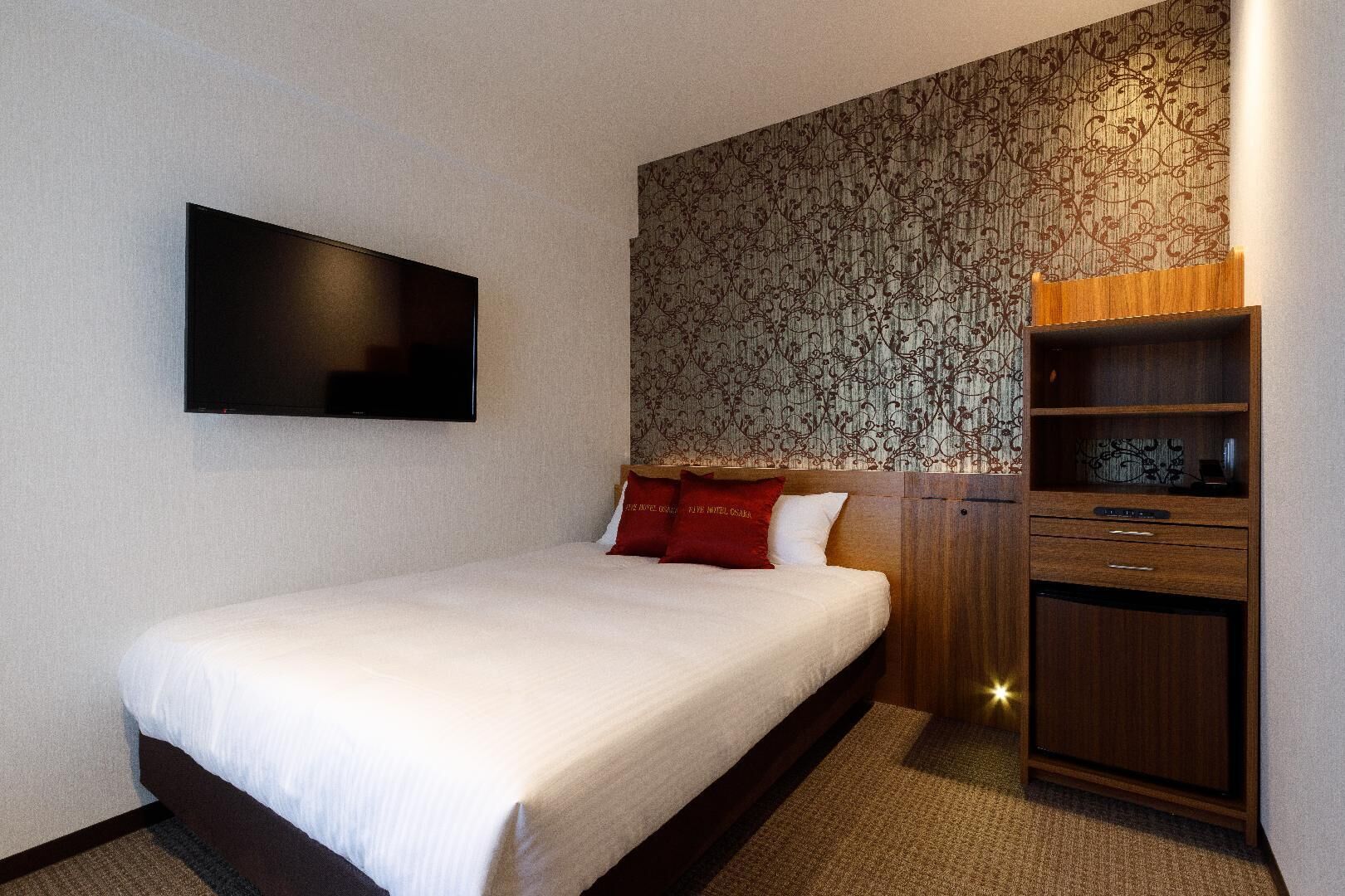 Quarto de hotel com cama, televisão na parede, mesa de cabeceira de madeira e papel de parede estampado.