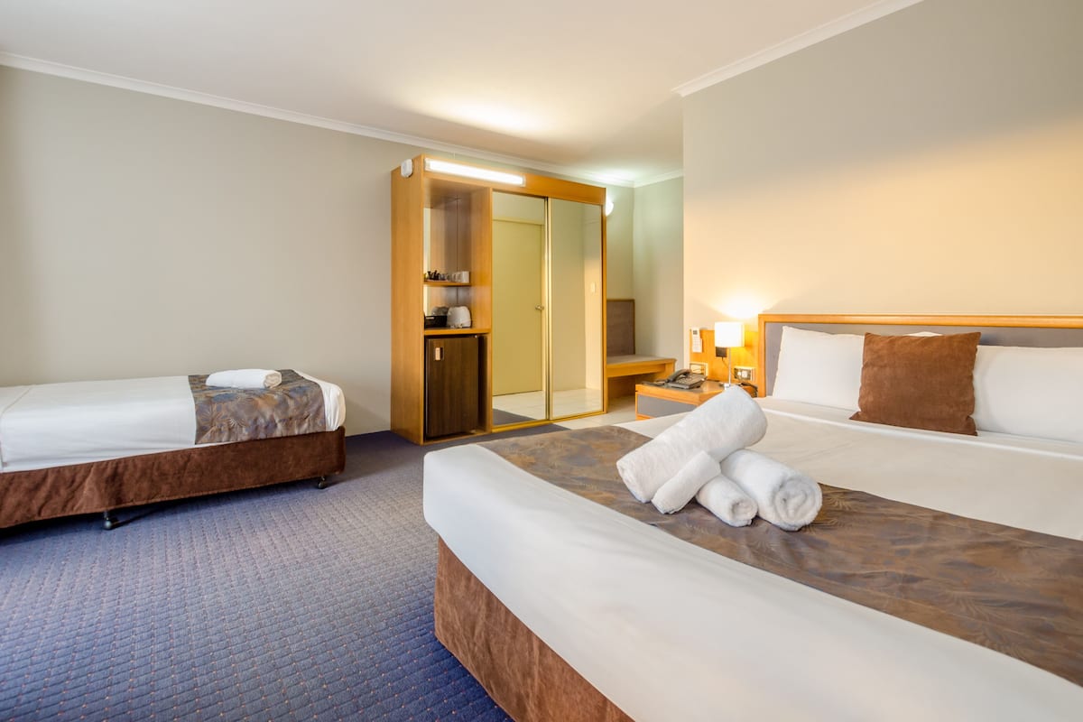 A hotel room with two beds, a wardrobe, a mirror, a bedside table with a lamp, and a bathroom visible through a glass door.