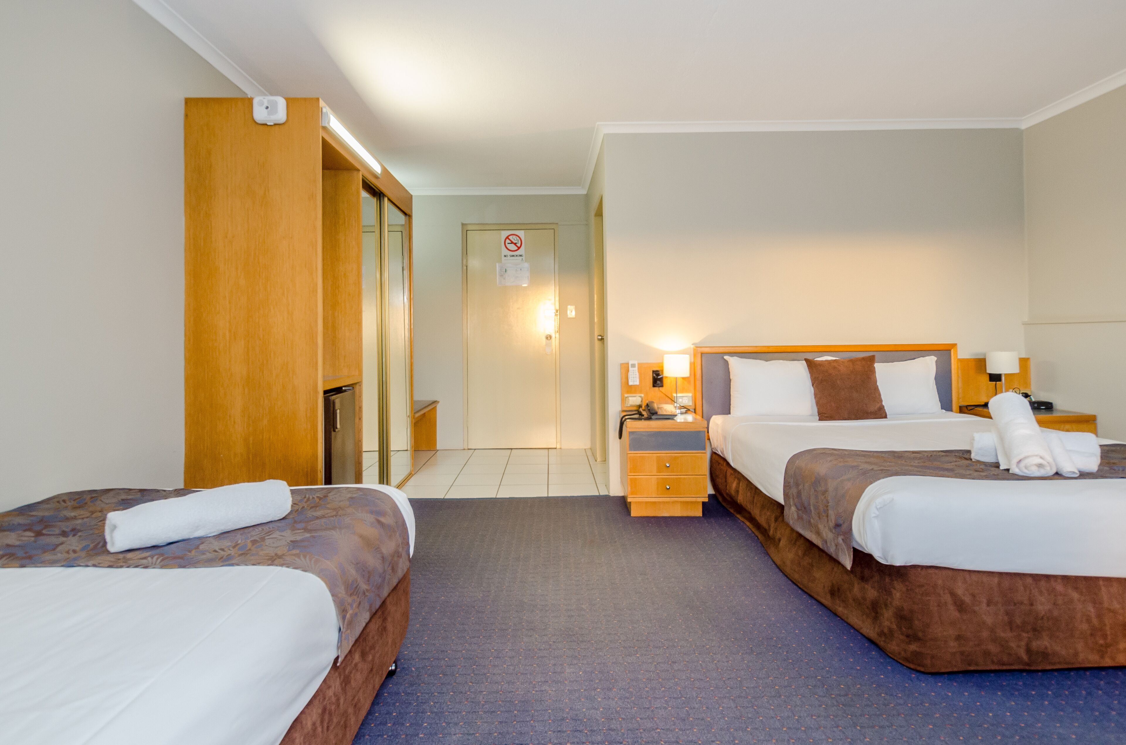 A hotel room with two beds, a wooden wardrobe, a desk with a lamp, and a bathroom door.