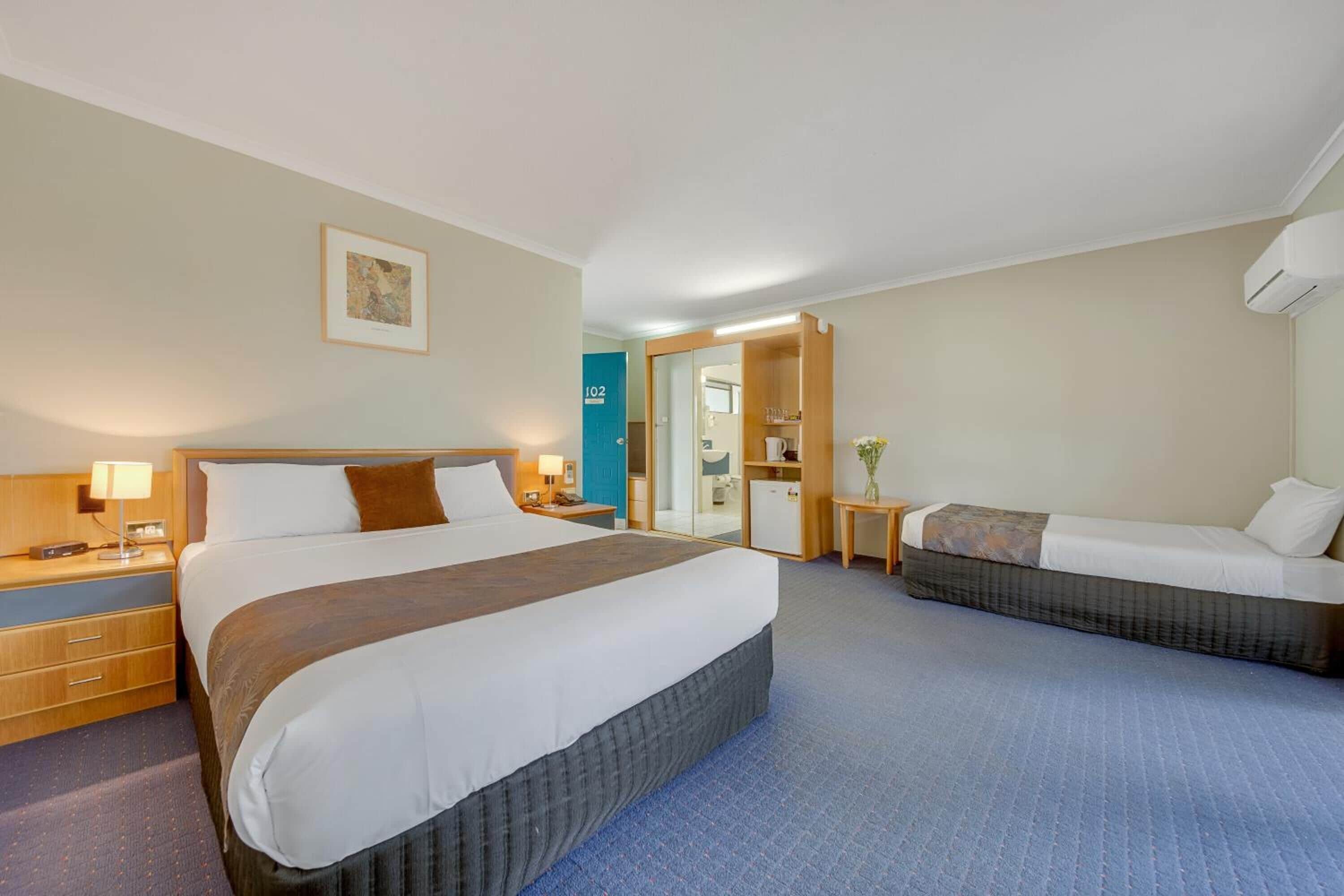 A hotel room with a large bed, a smaller bed, a desk, a chair, a nightstand, a lamp, and a mirror.