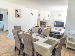 A dining area with wicker chairs, a table with a white tablecloth, a fireplace, a television, and a kitchen area in the background.