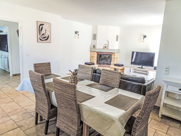 Un coin repas avec des chaises en osier, une table recouverte d’une nappe blanche, une cheminée, un téléviseur et, au fond, un espace cuisine.