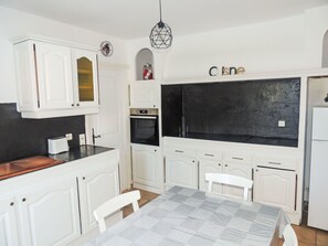 Une cuisine de taille réduite avec des meubles blancs, un pare-feu noir, une table à manger recouverte d’une nappe à motifs et un luminaire suspendu.