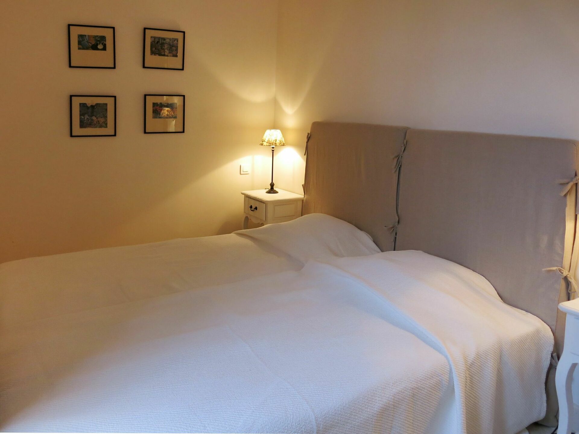 Un letto rifatto con un copriletto bianco e cuscini, un comodino con una lampada, e quattro quadri appesi al muro.