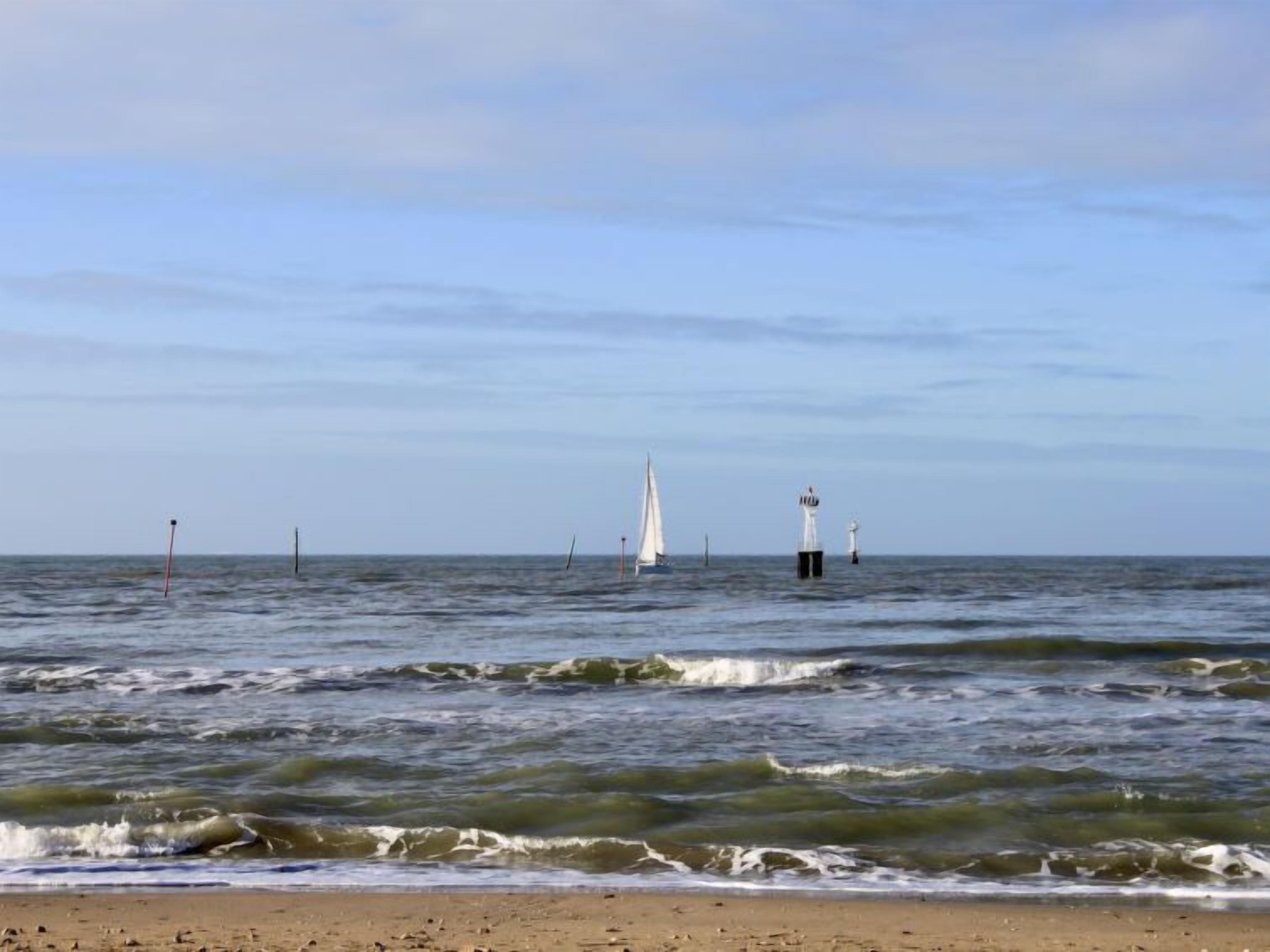 Ein Strand mit Wellen, einem Segelboot und einem Leuchtturm in der Ferne.
