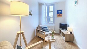 A compact living space with a sofa, a small table with a lamp, a TV, and a window with a view of buildings. - Apartment Carnot by Interhome (Deauville-Trouville)