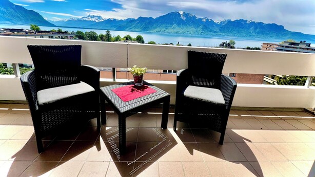A balcony with two wicker chairs and a small table, overlooking a mountainous landscape and a body of water.