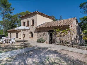 Un edificio de piedra con techo de tejas, un patio con mesas al aire libre y una pequeña zona ajardinada.