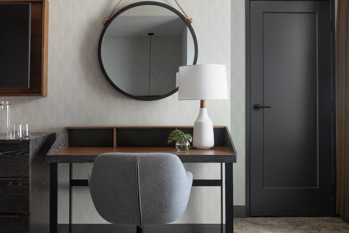 A modern hotel room with a wooden desk, a grey chair, a round mirror, a white lamp, and a door.