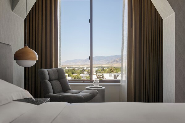 Una habitación de hotel moderna con un ventanal grande, un sillón gris, una mesita auxiliar pequeña y una lámpara colgante.