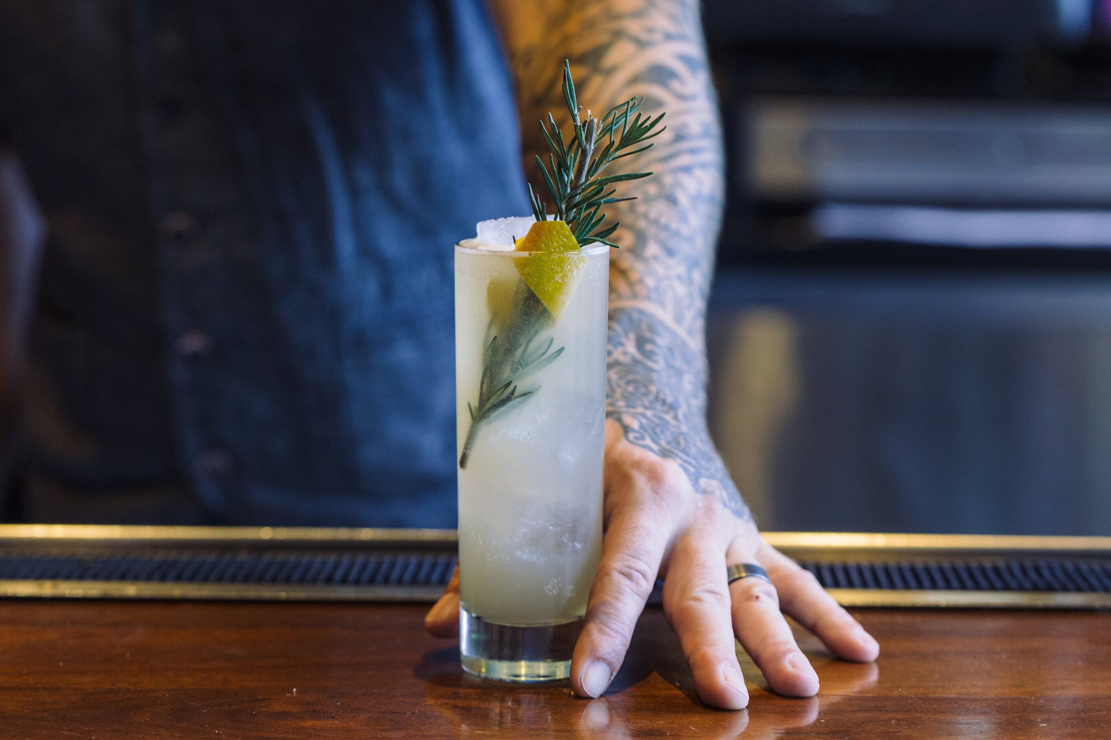 Drinkki, jossa on koriste, ja jonka kädessään pitää tatuoitu henkilö, baaritiskillä.