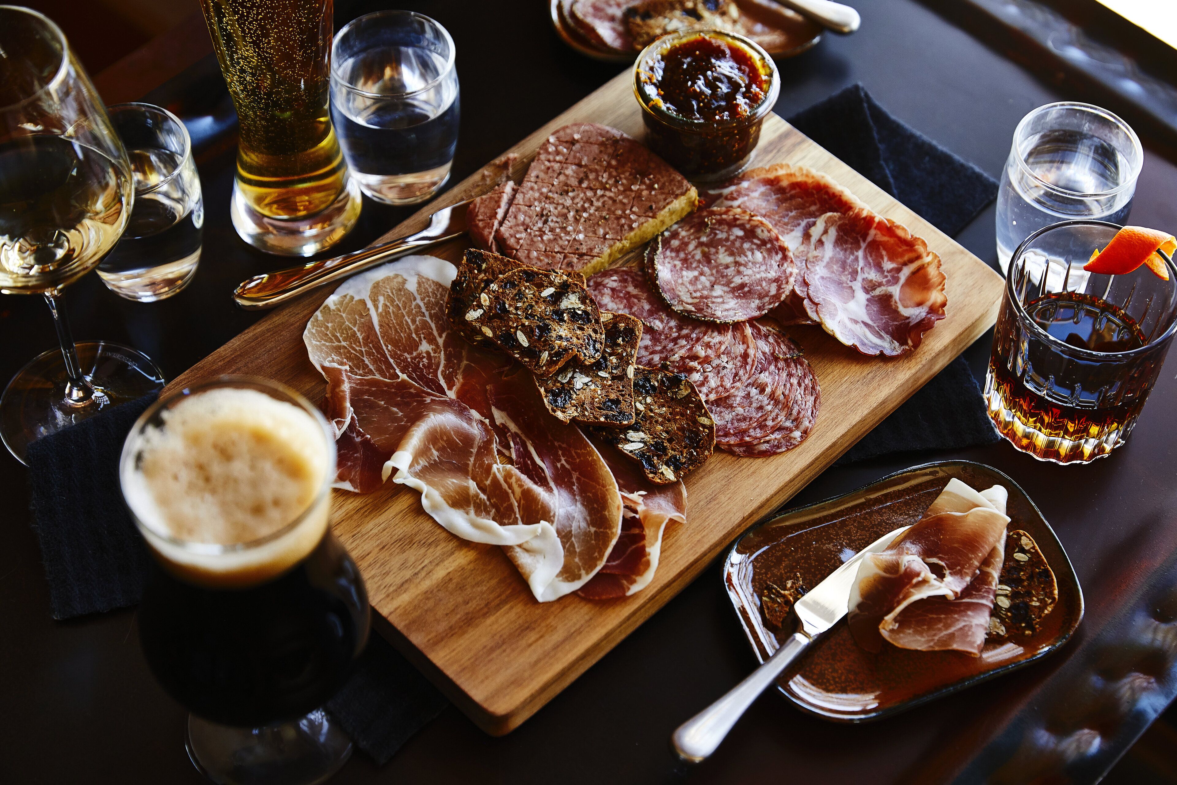 A wooden board with various cured meats, a glass of beer, and a bottle of water.